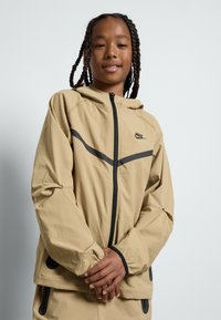 Casaco Nike de tom claro com capuz, apresentando detalhes em cinza escuro, fecho com zíper, punhos elásticos e bolsos laterais. Tecido de textura suave.