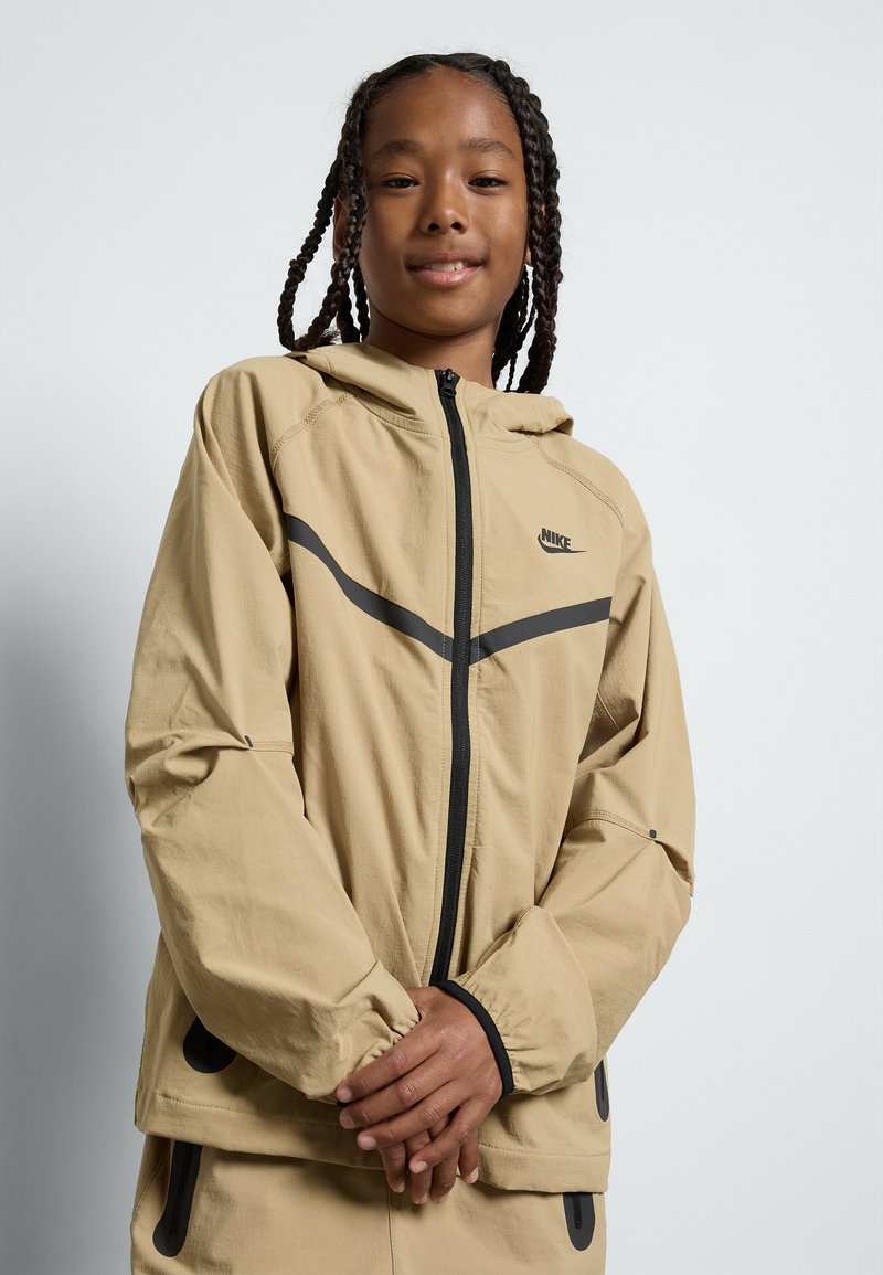 Casaco Nike de tom claro com capuz, apresentando detalhes em cinza escuro, fecho com zíper, punhos elásticos e bolsos laterais. Tecido de textura suave.