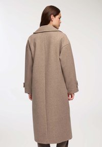Kobieta w długim beżowym wełnianym płaszczu z szerokim kołnierzem i paskami przy mankietach, stojąca tyłem do aparatu na jednolitym tle.