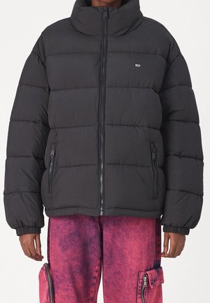 Zwarte gewatteerde pufferjas met hoge kraag en ritssluiting aan de voorkant, gedragen met roze acid-wash cargobroek met ritssluitingen op de zakken.