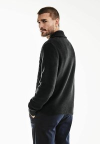 Suéter negro de punto con un acabado texturizado, que presenta un cuello chal y puños acanalados, combinado con pantalones oscuros. Modelo visto desde la espalda.