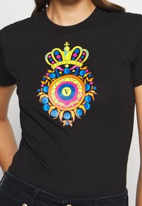 Zwart shirt met kleurrijk wapen dat een kroon, blauwe en roze bollen, en tekst "Versace Jeans Couture" in de centrale cirkel toont.