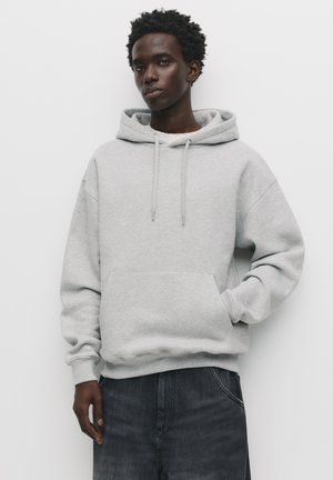 Light grey hoodie made of soft material, featuring a front pouch pocket, drawstrings, and ribbed cuffs. Worn with dark denim pants.