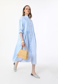 Robe rayée bleu clair avec un devant boutonné, des manches ballon et une taille froncée. Associée à un sac à main tressé beige et des ballerines grises.
