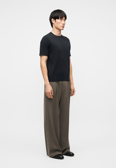 Άνδρας στέκεται στραμμένος προς τα δεξιά φορώντας μαύρο κοντομάνικο πουκάμισο, φαρδιά καφέ παντελόνια και μαύρα παπούτσια μπροστά σε απλό λευκό φόντο.