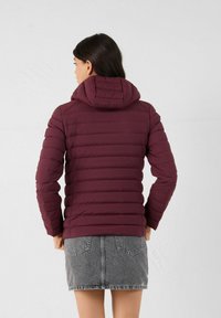 Giacca a vento bordeaux con un design aderente, texture trapuntata orizzontale e cappuccio. Indossata con una gonna di jeans grigia.