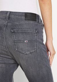 Jeans de mezclilla gris con una etiqueta de cuero oscura que dice "TOMMY JEANS" en la cinturilla, con un bolsillo trasero que presenta un pequeño logo en rojo, blanco y azul.