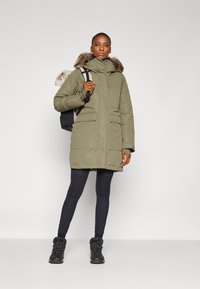 Groene parka met een met bont gevoerde capuchon, voorzien van twee voorzakken, gedragen over zwarte leggings en zwarte laarzen met gestructureerde veters.
