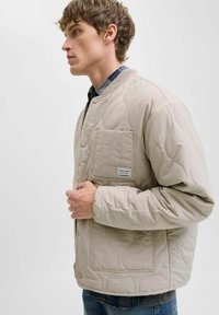Veste matelassée beige en matériau léger, comportant une poche à rabat, une fermeture à boutons-pression et un motif en vagues texturé.