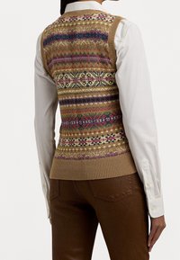 Personne portant un gilet tricoté à motifs colorés par-dessus une chemise blanche à manches longues et un pantalon marron, vue de dos.
