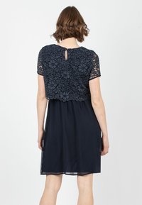 Navy Kleid mit einer Spitzenoberfläche, kurzen Ärmeln und einem runden Ausschnitt. Der Rücken hat ein kleines Schlüssellochdetail und einen fließenden Rock.