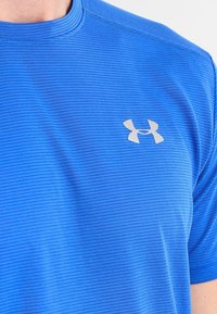 Camisa atlética azul de manga corta con un patrón de rayas texturadas y un pequeño logo plateado de Under Armour en el lado izquierdo del pecho.