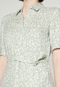 Vestido de manga curta em tecido verde claro com padrão abstrato branco, apresentando uma frente com botões e um detalhe de cintura atada.