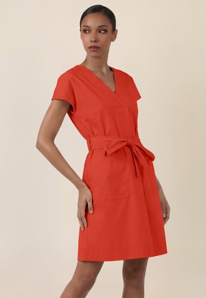 Woman with tied waist red dress, short sleeves, V-neck, standing with one hand on hip, looking to side against plain background.