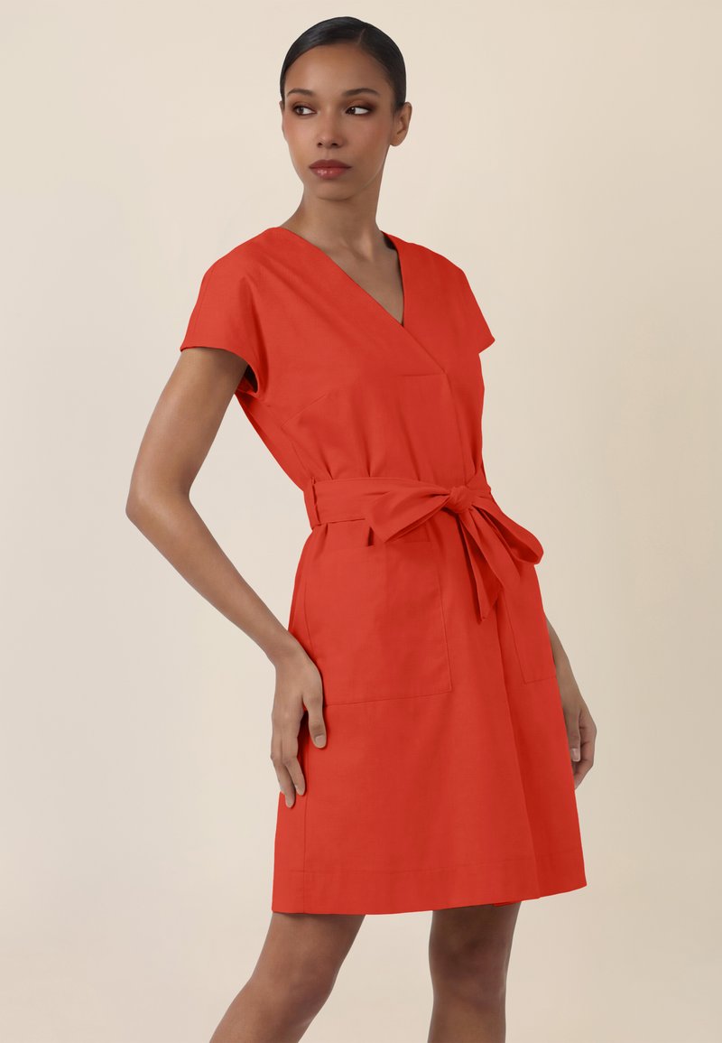 Femme portant une robe rouge à taille ceinturée, manches courtes, encolure en V, debout avec une main sur la hanche, regardant de côté, sur un fond uni.