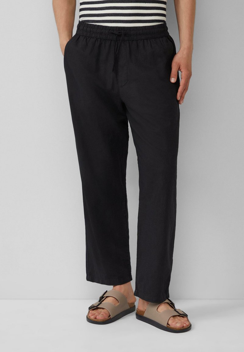 Pantalons noirs, légers et amples avec une taille élastique et un cordon de serrage. Associés à des sandales marron avec une détail de boucle.