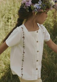 Camicia bianca a maniche corte con scollatura quadrata, caratterizzata da ricami floreali lungo il lato e bottoni in legno lungo la parte anteriore.