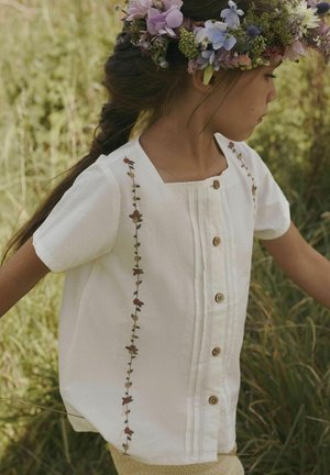 Camicia bianca a maniche corte con scollatura quadrata, caratterizzata da ricami floreali lungo il lato e bottoni in legno lungo la parte anteriore.