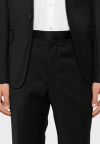 Giacca nera con un solo bottone, abbinata a pantaloni neri. Il tessuto appare liscio con una sottile texture. La camicia ha dettagli dei bottoni visibili.