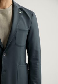 Sacou verde închis cu rever, închidere cu un singur nasture și buzunar tip patch. Textură de țesătură netedă, cu croi slim și design minimalist.
