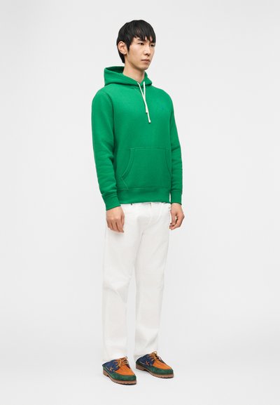 Mladi moški stoji, oblečen v zeleno kapucarico, bele hlače in večbarvne rjave, modre ter zelene čevlje na enakomerni belem ozadju.