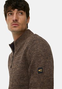 Brauner, gestrickter Pullover mit Reißverschluss und hohem Kragen. Verfügt über ein strukturiertes Muster und einen schwarzen Patch mit goldener Logoschrift auf dem linken Ärmel.