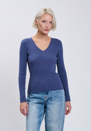 Donna con capelli biondi corti che indossa un maglione blu a coste con maniche lunghe e scollo a V e jeans azzurri chiari, in piedi davanti a uno sfondo chiaro e uniforme.