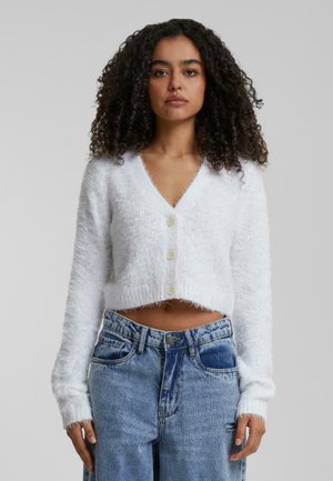 Hvit kort cardigan laget av fluffy materiale, med V-hals, lange ermer og fire frontknapper. Sammen med høyhalsede blå jeans.