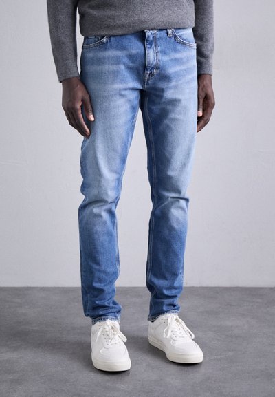 Jean en denim bleu clair avec une coupe slim, zones décolorées et design à cinq poches, associé à des chaussures de sport blanches et un pull gris.