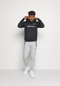 Schwarze Windjacke mit Kapuze und Logo, kombiniert mit hellgrauen Jogginghosen. Verfügt über elastische Bündchen und Taschen. Weiße Sneakers runden das Outfit ab.
