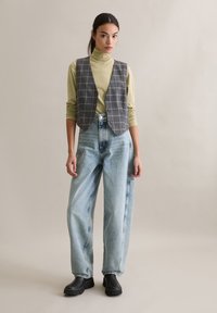 Szara kamizelka w kratę na jasnozielonym golfie, zestawiona z luźnymi niebieskimi dżinsami i czarnymi chodakami. Gładkie tekstury i luźny krój.
