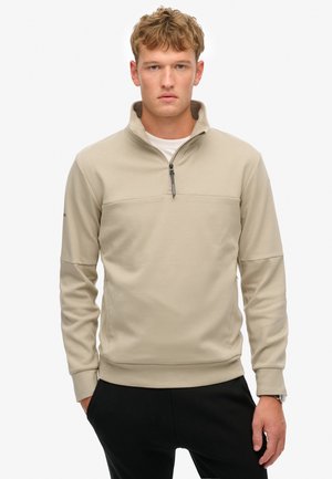 Sudadera beige de media cremallera, hecha de un tejido suave, con un panel frontal texturizado y puños acanalados, combinada con pantalones negros.