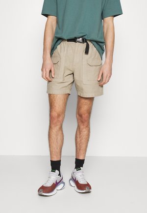 Beige cargoshorts met twee voorzakken, elastische tailleband en riemlussen, gecombineerd met een groene oversized T-shirt en meerkleurige sneakers.