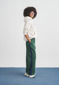 Sweatshirt bege claro com bolso frontal, combinado com calças verdes com risca azul-marinho. Modelo usa óculos e ténis brancos num chão azul.