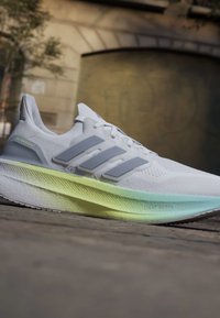 Chaussure de running blanche Adidas Ultraboost avec des rayures grises et une semelle en dégradé vert clair à bleu sur un revêtement extérieur.