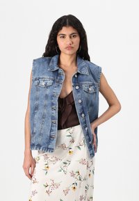 Jeansväst utan ärmar i ljusblått, med klassisk krage, framfickor och knäppning med knappar. Stylad över en brun topp och blomsterkjol.