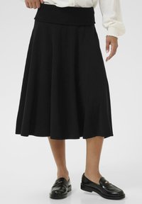 Jupe évasée noire avec une large ceinture côtelée, présentant une texture lisse et un ourlet au niveau des genoux. Associée à des mocassins noirs brillants.
