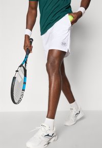 Hvite tennis shorts med EA7-logo, grønn treningsskjorte, holder en blå og svart tennisracket, iført hvite sneakers og sokker.