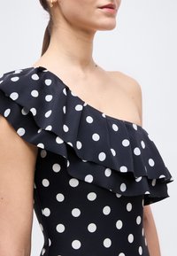 Γυναίκα που φοράει μαύρο φόρεμα με έναν ώμο, με λευκές πουά και στρωμένο βολάν, με εμφανές χρυσό κρίκο στο αυτί.