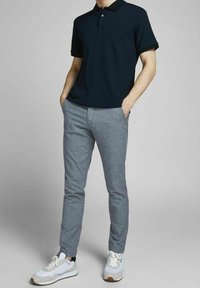 Mężczyzna w czarnej koszulce polo z krótkim rękawem, szarych dopasowanych spodniach i białych sportowych trampkach, z rękami w kieszeniach.