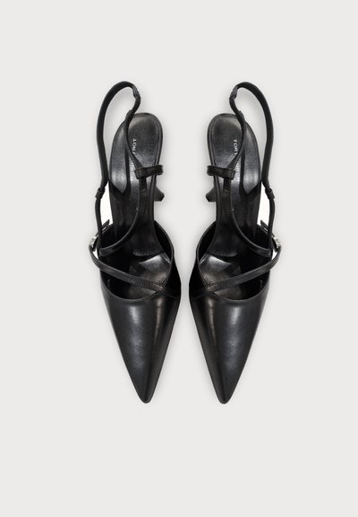 Μαύρες γόβες slingback με μυτερή μύτη, λεπτούς λουράκια που διασταυρώνονται και χαμηλά τακούνια kitten σε ανοιχτό φόντο.