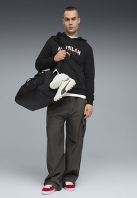 Sweat à capuche noir avec logo "AC MILAN", pantalon cargo gris, baskets rouges stylées, portant un sac de sport noir avec des chaussures de football blanches attachées.