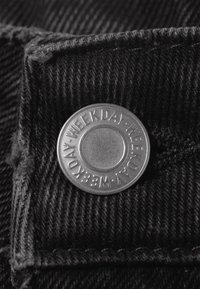 Bouton en métal sur un tissu texturé foncé avec le mot "WEEKDAY" en relief répété autour de son bord.