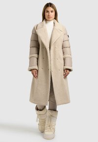 Beige lange jas met een gestructureerde, pluizige buitenkant en gevoerde mouwen. Heeft een ruime kraag, dubbele knoopsluiting en zijzakken.