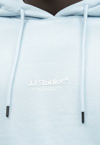 Jasnoniebieska bluza z kapturem z przednią kieszenią, ściągaczami z czarnymi końcówkami oraz logo "JJ Studios" nadrukowanym na białym tle na piersi.