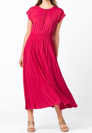 Femme portant une robe midi fluide rose vif avec une taille froncée et des manches courtes, associée à des talons ouverts beige, debout sur fond blanc.