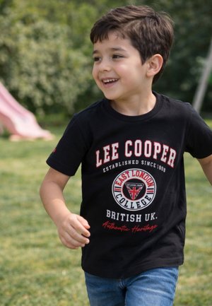 Garçon souriant portant un T-shirt noir Lee Cooper et un jean bleu, courant en plein air sur l'herbe avec des arbres et un toboggan rose en arrière-plan.