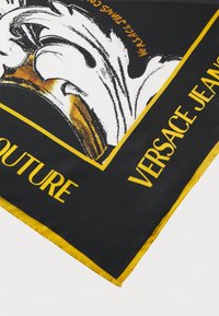 Musta silkkihuivi, jossa on rohkea valkoinen ja kulta grafiikkasuunnittelu, keltainen reuna ja "Versace Jeans Couture" -teksti alareunassa.