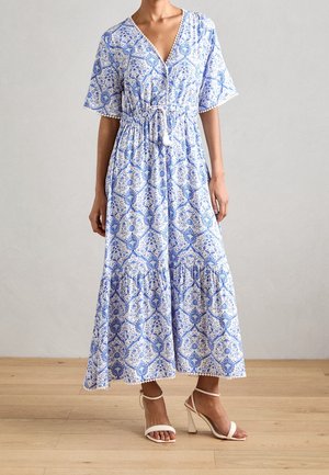 Vrouw draagt een midi-jurk met blauw-wit patroon, korte mouwen en een V-hals, gecombineerd met witte sandalen met hoge hakken.