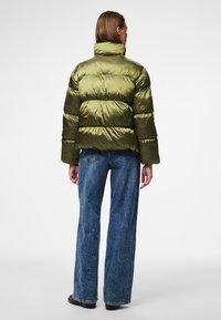 Olive green puffer jacket made of shiny material, featuring horizontal quilted pattern, high collar, and a loose fit. Paired with flared jeans.
